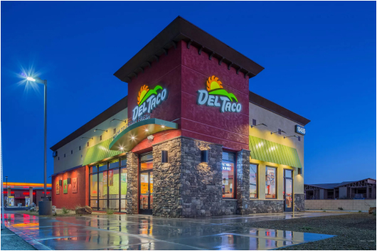 Angled exterior photo of a Del Taco restaurant at night. Signage above both sides of the entrance shows the restaurant logo of a sun coming over the mountains and “Del Taco” in text below it.